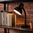 Lloytron 35W Classic Flexi desk lamp, black, lit on a bookshelf against a brick wall