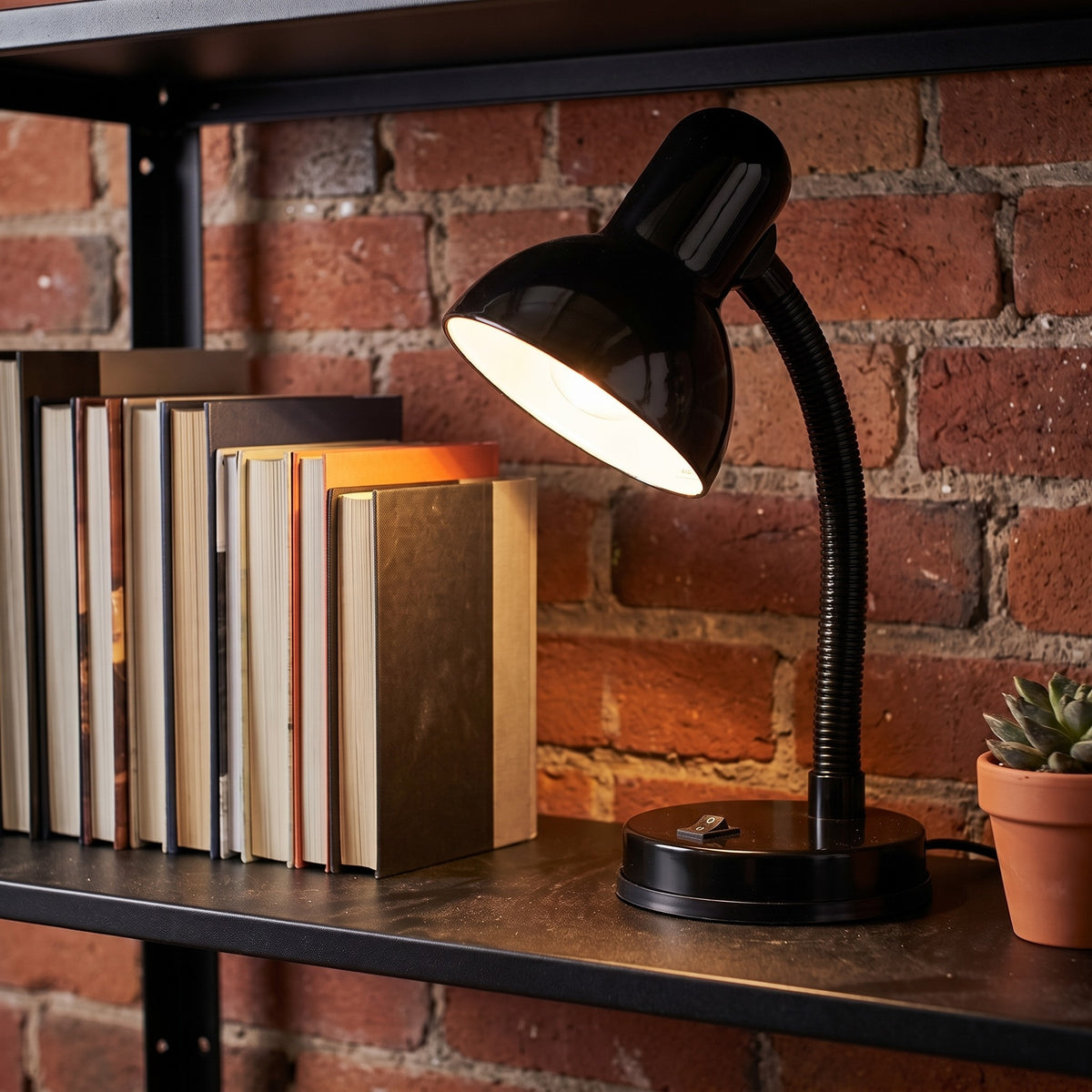 Lloytron 35W Classic Flexi desk lamp, black, lit on a bookshelf against a brick wall